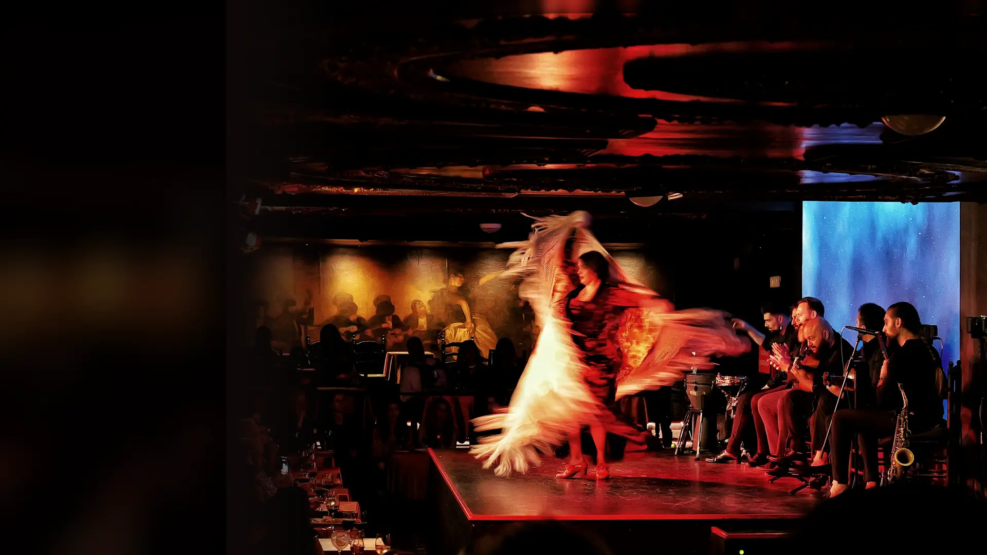 A flamenco dancer in a swirling red and cream shawl and skirt turns on a lit stage while seated musicians play guitars and a saxophone behind her and diners watch from tables in the dark room