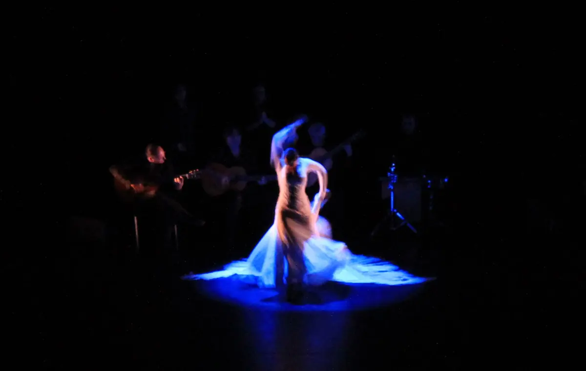 A flamenco dancer in a flowing blue dress turns on a dark stage lit in blue, with musicians seated behind her