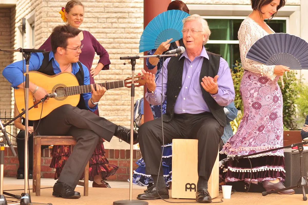 A flamenco singer seated on a cajon, a guitarist and two dancers holding blue fans perform together on an open-air stage