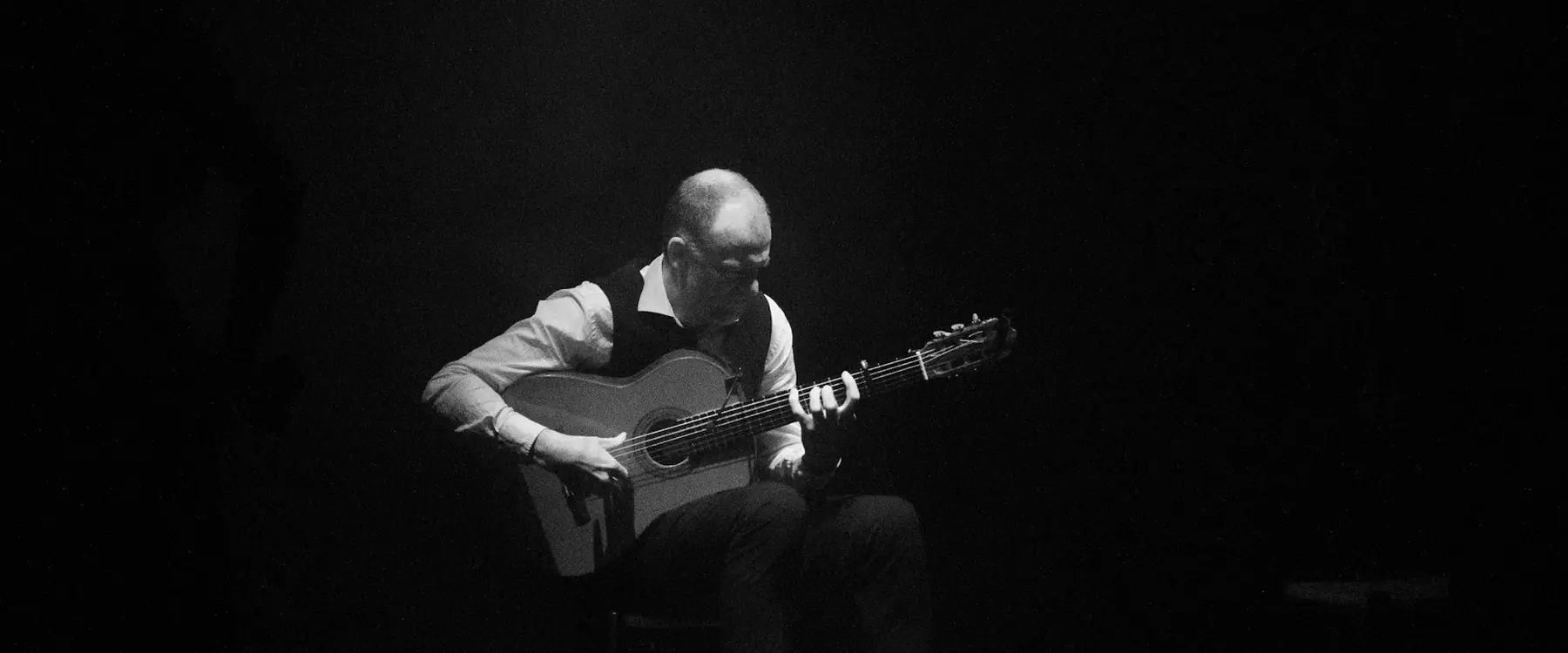 A guitarist seated in a single shaft of light in a black-and-white photograph, the rest of the stage in darkness