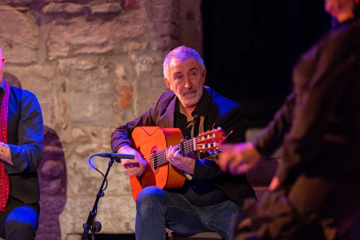 A flamenco guitarist in a dark jacket plays a golden-orange guitar on a small stone-walled stage, with a microphone in front of him and another performer at his shoulder
