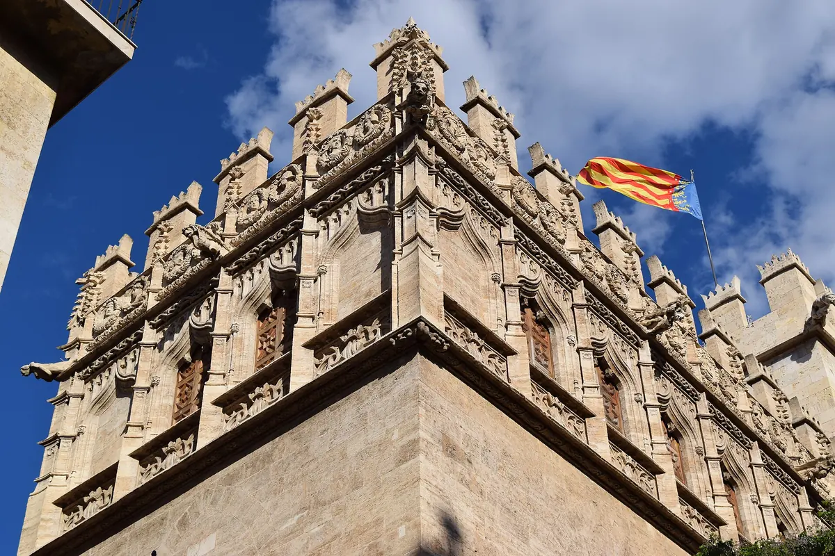 The crenellated stone corner of the Silk Exchange in Valencia, with carved windows and a flag against a blue sky