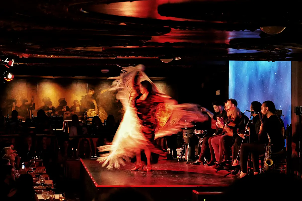 A tablao dining room in red light, with a raised stage where a dancer turns in a swirling shawl, seated musicians behind her and diners at tables in the foreground