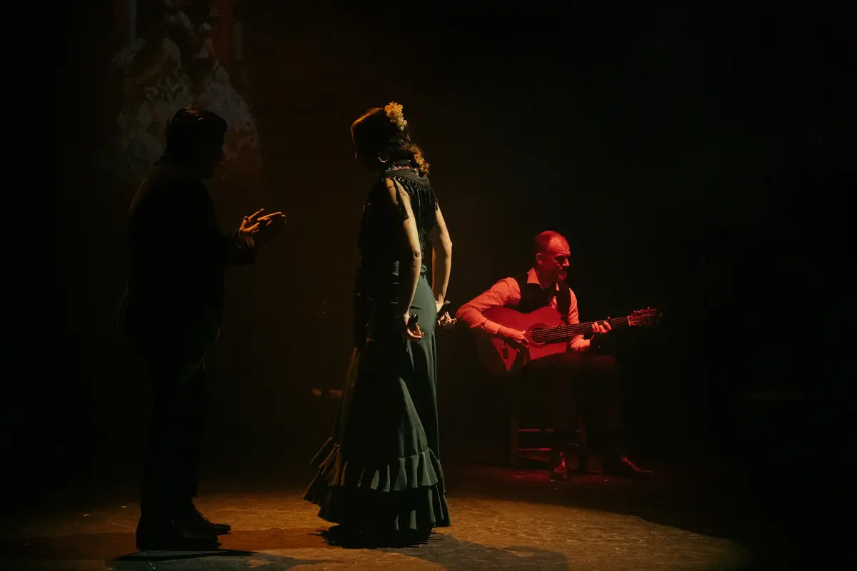 A dancer in a long dark ruffled dress stands in a pool of amber light beside a man in silhouette, while a guitarist sits at the right edge of the stage